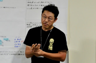 左胸に「南三陸」と書かれた黒いTシャツを着た男性教師が、模造紙の前で話している様子をアップで撮影した写真