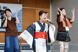 浴衣姿の女性がマイクで歌いながら、両脇のスタッフが手拍子に合わせて笑顔で踊っている様子が写っている写真