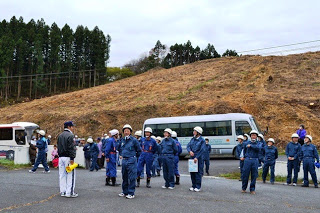 青い防災服と白いヘルメットを着用した多くの参加者がバスの前で集合している様子の写真