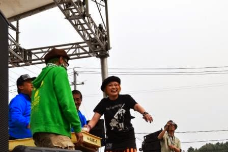 ステージの上で笑顔のまちゃまちゃさんが観客に向かって、ばんじゅうに入った餅を観客に向かって撒こうとしている様子の写真