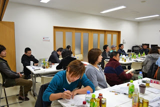 ペットボトルや資料が並ぶテーブルで、ノートにメモを取りながら集中して講義を受ける参加者たちが座っている写真