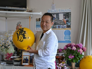 花瓶に生けられた花や写真、ポスターが貼られた室内で、白い服を着た男性が「慶」と筆書きされた黄色い球体を笑顔で持っている写真