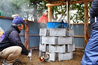 組み立てられた簡易炭焼き炉に火が入り、男性が側で炎の様子を確認している様子を写した、炭焼きの点火直後の写真