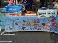様々な魚などのイラストが描かれた横断幕をテーブルの下に設置した屋台に、こんぶやわかめなどの海産物が並んでいる写真