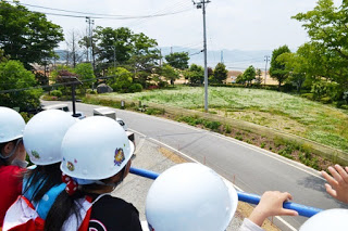 バケットに乗ったヘルメットをかぶった園児たちが、道路や草原、木々の先に湾が見える景色を見ている写真