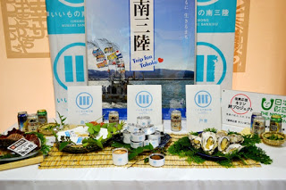 ーブルの上には缶詰、牡蠣、海産物の盛り合わせ、地元の加工食品やパンフレットが並べられ、背景には「南三陸」や「Trip for Tohoku」と書かれた大きなパネルが立てられている、三陸の観光や特産品をPRする展示ブースの写真