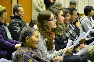 観客席で地元の高齢者や外国人、若者たちが一緒に歌詞カードを持ちながら合唱に参加している音楽交流の写真