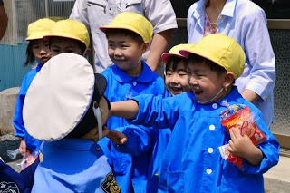 青いスモッグを着て黄色の帽子をかぶった5人の園児が、警察の制服と帽子を身に着けた園児を笑顔で見ている写真