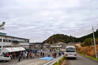 山あいの地域にある建物前の広場で多くの人々がテントの下や建物周辺に集まり黙祷を行っている様子の写真