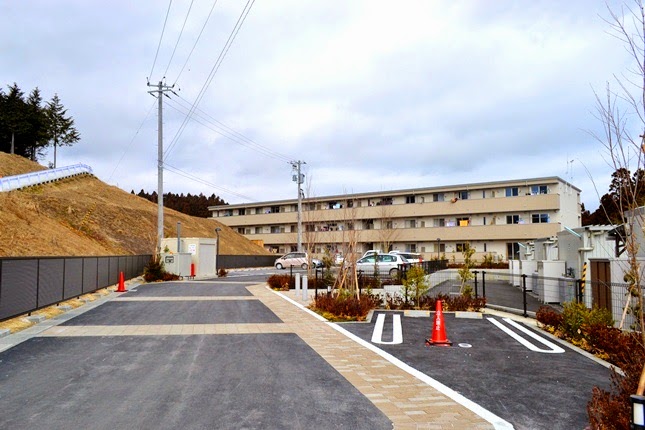 整備された舗装道路の奥に建つ中層の集合住宅と、その周囲に植栽や駐車スペースが整えられた新興住宅地の写真