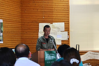 緑色の作業服を着た男性が演台に立って聴衆に向かって話している様子がアップで写っているセミナーの写真