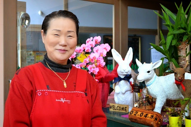 白いうさぎとやぎの置物のそばで、赤い服とエプロンを着た笑顔の及川福子さんが写っている写真