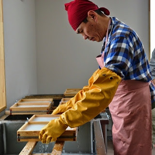 赤いバンダナを頭に巻き、エプロンに長い手袋をつけた男性が、紙漉きを行っている様子を横から撮影した写真