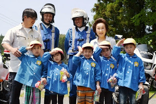 白バイ隊員の青い制服とヘルメットを着用した男性2人と関係者の男女の前に並んで立ち敬礼のポーズをしている、青いスモッグと黄色い帽子を身に着けた5人の園児の写真