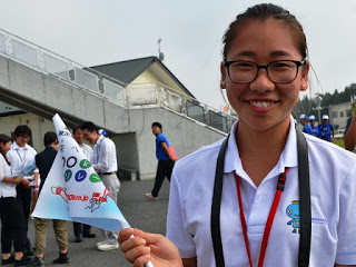 陸橋の近くで小さな旗をもった亀山さんが笑顔でカメラをみている写真