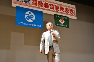 「南三陸町高齢者芸能発表会」と書かれた横断幕が掲げられたステージで、右手にマイクを持った司会の猪又事務局長が話をしている写真