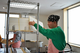 黒色のバンダナを頭に巻き、ピンク色のエプロンを着用した男性が、ステンレス製のラックの上段に漉いた紙を立てかけて乾かしている様子の写真