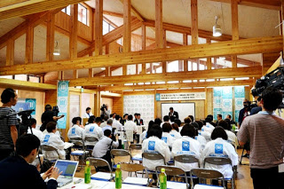 木造の建物の中で、多くの人々が椅子に座って前方の話し手に注目している様子が写っている協議会の様子の写真