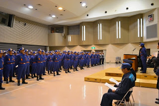 青い制服と帽子を着た大勢の消防団員が前を向いて整列し、壇上では指揮官らしき人物が話している写真