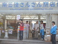 南三陸さんさん商店街と屋根に書かれた大きなテントの前に5人の男性が立っており、左端の男性がマイクを持ってい話をしている様子の写真