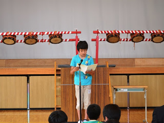 他の児童が床に座って聞いているなか、水色のシャツを着た男の子が演台の前に立ち、大きな紙を読み上げながら発表している写真