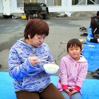 防災訓練の会場で青いレジャーシートに座り、豚汁を食べている母親と、隣に恥ずかしそうに座っている女の子の写真