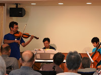 バイオリン・ピアノ・チェロを演奏する人々が前方におり、その演奏を聴く観客が後方に座っている様子を写した写真