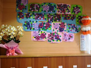 木製の壁に紫や赤、緑の折り紙で作った花を青やピンクの養子に貼りつけて作ったアジサイの作品がたくさん展示されており、左側にはピンクの包装に包まれた白い蘭が置かれている写真