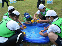 芝生の上に置かれた小さな青いビニールプールの周りで、緑色のビブスと白い帽子を着た男性4人と子供一人がが水風船を作っている様子を写した写真