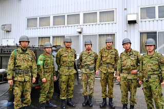 白い建物の前に設置された炊飯用トレーラーで炊き出しを行っているヘルメットをかぶった自衛隊員7人が、整列し真っ直ぐ前を見て立っている写真