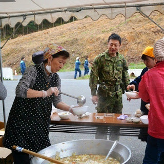 テントの下で女性が大きな鍋からお玉で豚汁をよそい、自衛隊員を含む参加者に配膳している様子の写真