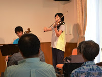 部屋の中で数人の観客が座っている前でバイオリンを弾いている女性とチェロを弾いている男性が演奏している様子を写した写真