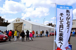 「ありがとう！そして共に未来へ！」と書かれたのぼり旗が手前にあり、その奥で人々が白いテントに向かって歩いている写真