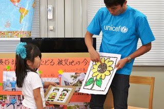 ひまわりの絵を持った長谷部選手が種の部分を指さし目の前の女の子に説明をしている写真