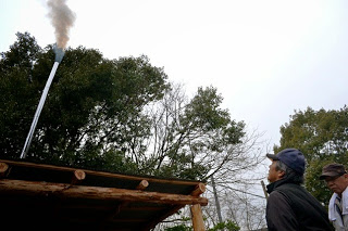 炉に取り付けた煙突から白い煙が立ち上り、屋根の外でそれを数人が見上げている様子を写した、炭化反応中の排煙の確認の写真