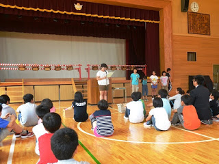 後ろで4人の児童が並び、他の児童たちが体育館の中央に座って聞いているなか、舞台の前で一人の女の子が発表している写真