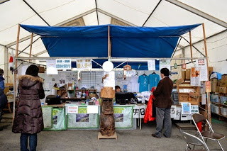 白いテントの中で、中央に木製のモアイ像が置かれ、物品や掲示が並ぶ青い布で屋根が作られた受付カウンターに数人の来訪者が並んでいる様子の写真