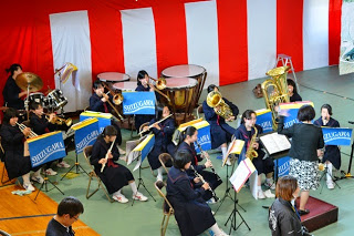 紅白幕がかけられた体育館内で、女性教師の指揮に合わせて、生徒たちが様々な楽器を演奏している吹奏楽部の様子を2階から撮影した写真