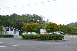 木々に囲まれた広い駐車場に車が数台停まり、白い建物の平成の森管理事務所の写真