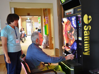 高齢の男性が室内に設置された黒いパチンコの台で楽しんでいる様子を女性スタッフが後ろからみている写真