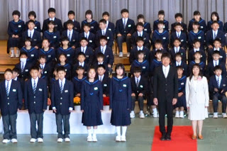 ステージ上と下に新しい制服に袖をとおした新入生が座り、手前にブレザーを着た3人の男子生徒と、セーラー服を着た2人の女子生徒、男性教師と女性教師が横一列に並んで立っている写真