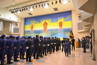 壁に大きなカラフルな消防士の壁画があるホールで、青と黒の制服を着た消防団員たちが整列し、式典が行われている様子を後方から撮影した写真