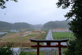 高台の鳥居の下から、まっすぐに伸びる道路と、整地された広い場所にシャベルカーが置かれた工事中の写真