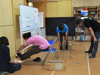 壁に向かってパイプ椅子に座ったピンクのシャツの人が両手を前に伸ばして体力測定をしている様子を写した写真