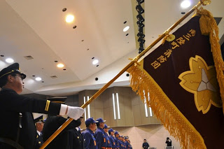 屋内で「南三陸町消防団」と金文字で書かれた大きな団旗を掲げる制服姿の男性の横に、青い制服の団員たちが整列している出初め式の写真