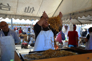 テントの下のブースで、白いエプロンを着た男性が、屋台の鉄板で焼きそばを大きなヘラで高く持ち上げながら調理している写真