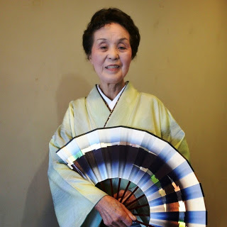 大きな扇を持ちながら薄い黄色の着物を着た女性が立っている写真