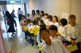 白いシャツを着た子どもたちが黄色い紙を持って舞台裏の廊下に並んで出番を待っている様子の写真