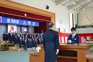 「第68回入学式」と書かれた横断幕が掲げられたステージに新入生が立ち、体育館の左側に設置された檀上にモーニングを着た校長先生と、新入生代表の男子生徒が向かい合って立ち誓いの言葉を述べている様子の写真