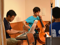 室内で3人の男性がそれぞれキーボード・チェロ・バイオリンを演奏している様子を横から写した写真